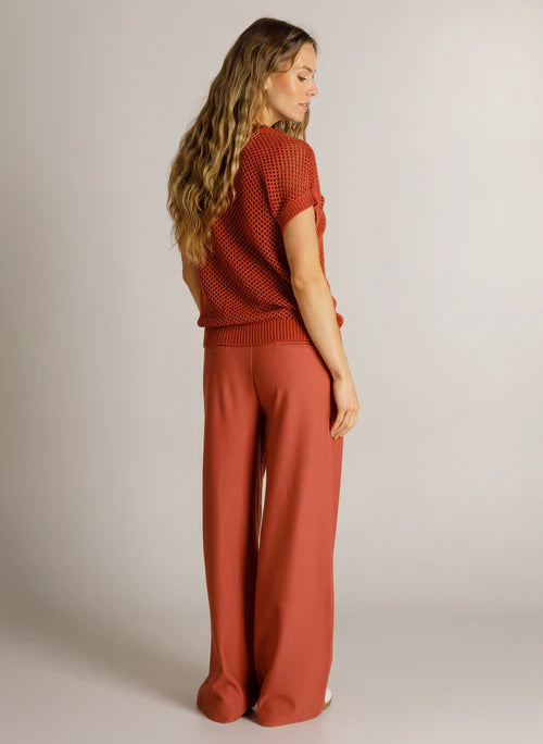 A woman wearing a rust-orange knit short-sleeve top and matching wide-leg pants, shown from the back in a studio setting with a light gray background.