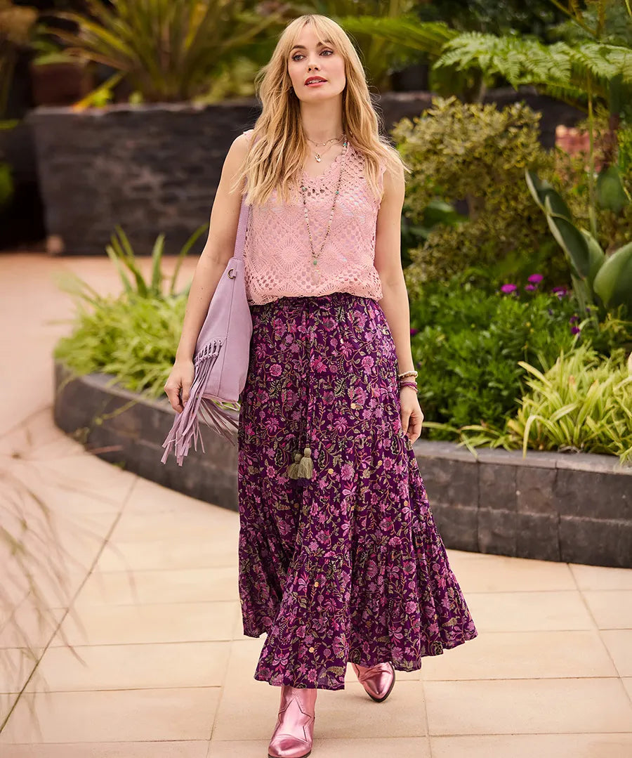 Woman in a pink top and purple floral skirt standing outdoors with plants in the background