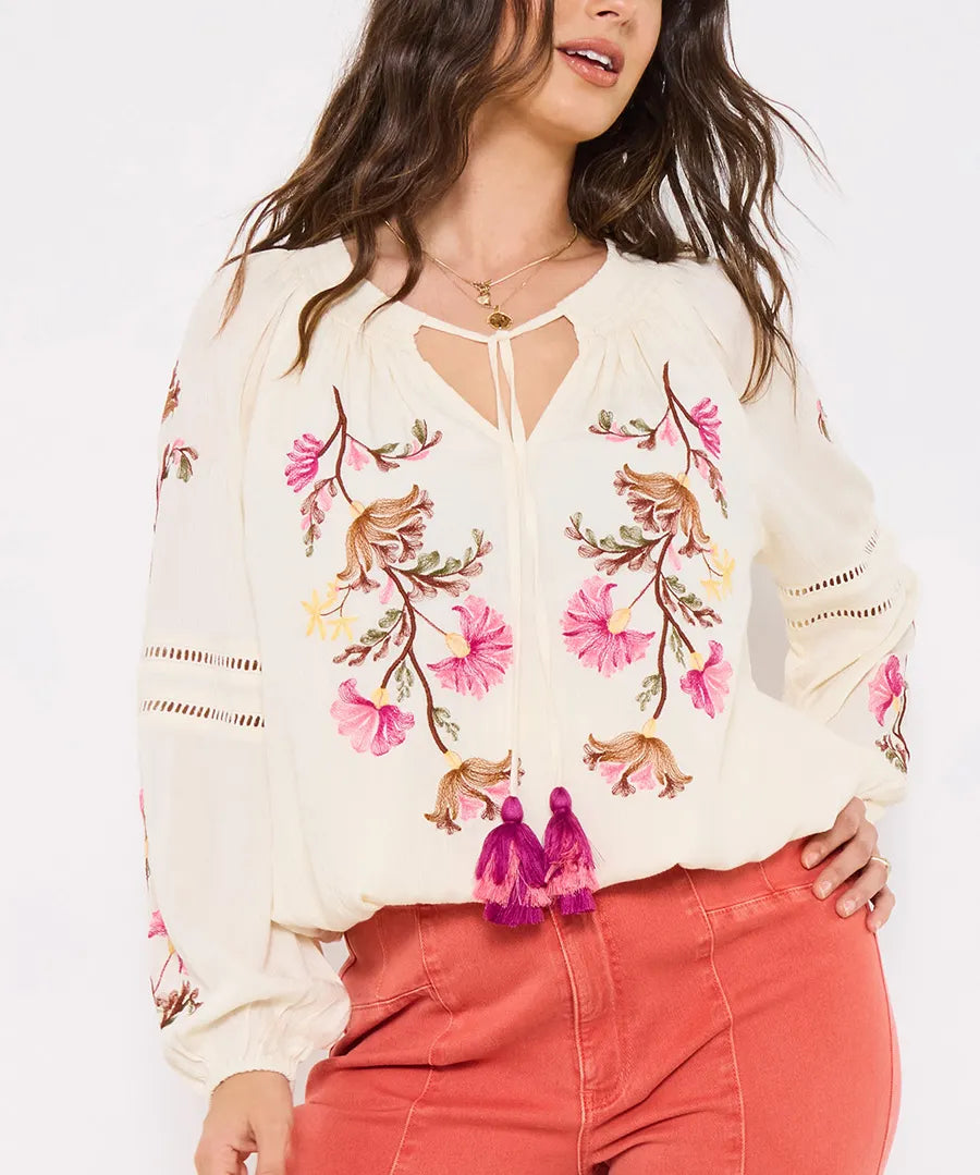 Woman wearing a white blouse with floral embroidery and tassels on a plain background