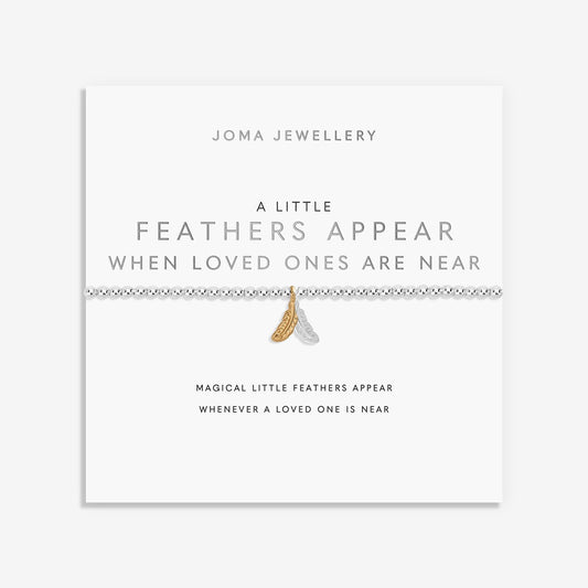 A delicate bracelet displayed on a white Joma Jewellery card. The bracelet features a silver-toned beaded line with a small gold-toned feather charm and a tiny white feather charm.
