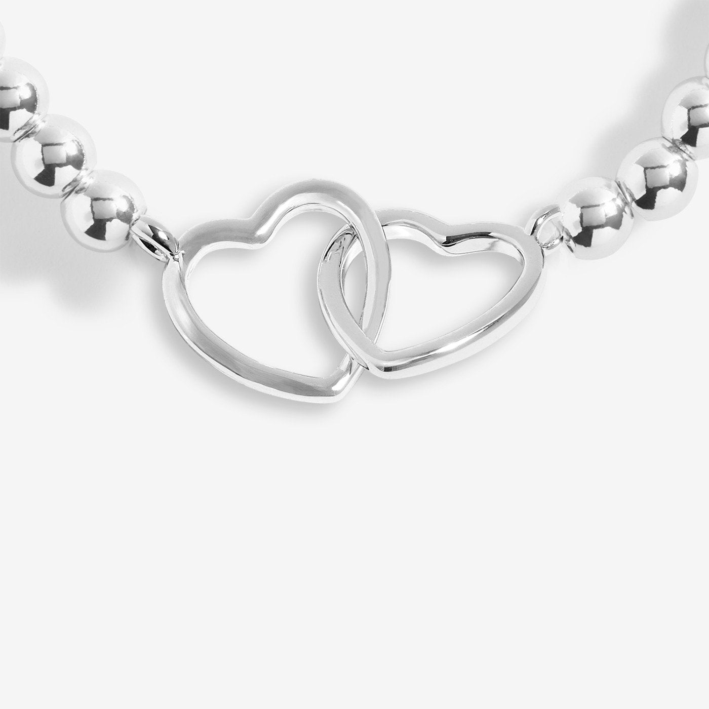 A close-up of a polished silver beaded necklace featuring two interlocking open heart charms at the center, set against a clean light gray background.