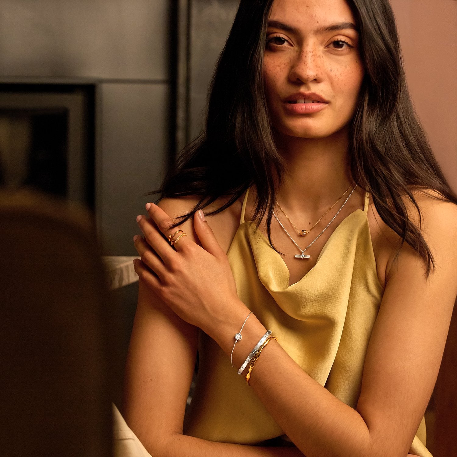 A woman wearing delicate silver jewelry, including a necklace and bracelets, paired with a yellow satin top in a warm-toned indoor setting.
