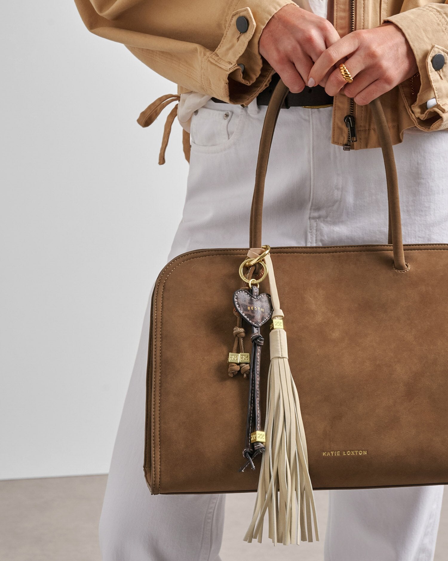 Brown leather handbag with tassel and keychain held by a person wearing a beige jacket and white pants.