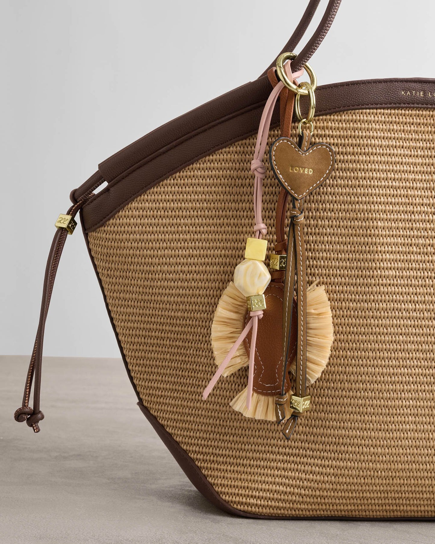 Brown woven handbag with decorative tassels and heart-shaped charm on a light gray background