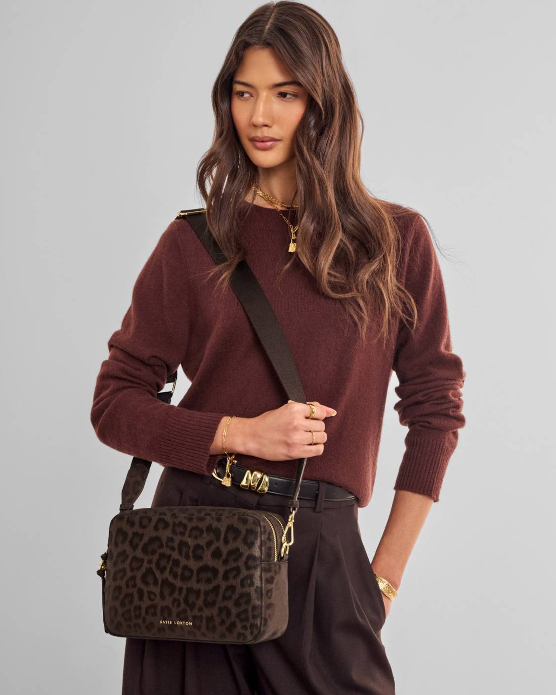 A woman wearing a brown leopard-print crossbody bag with gold hardware, styled with a burgundy knit sweater and dark brown trousers against a solid light gray background.