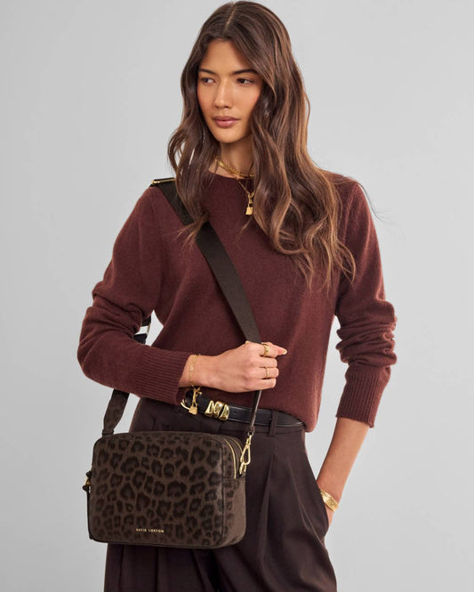 A woman wearing a brown leopard-print crossbody bag with gold hardware, styled with a burgundy knit sweater and dark brown trousers against a solid light gray background.