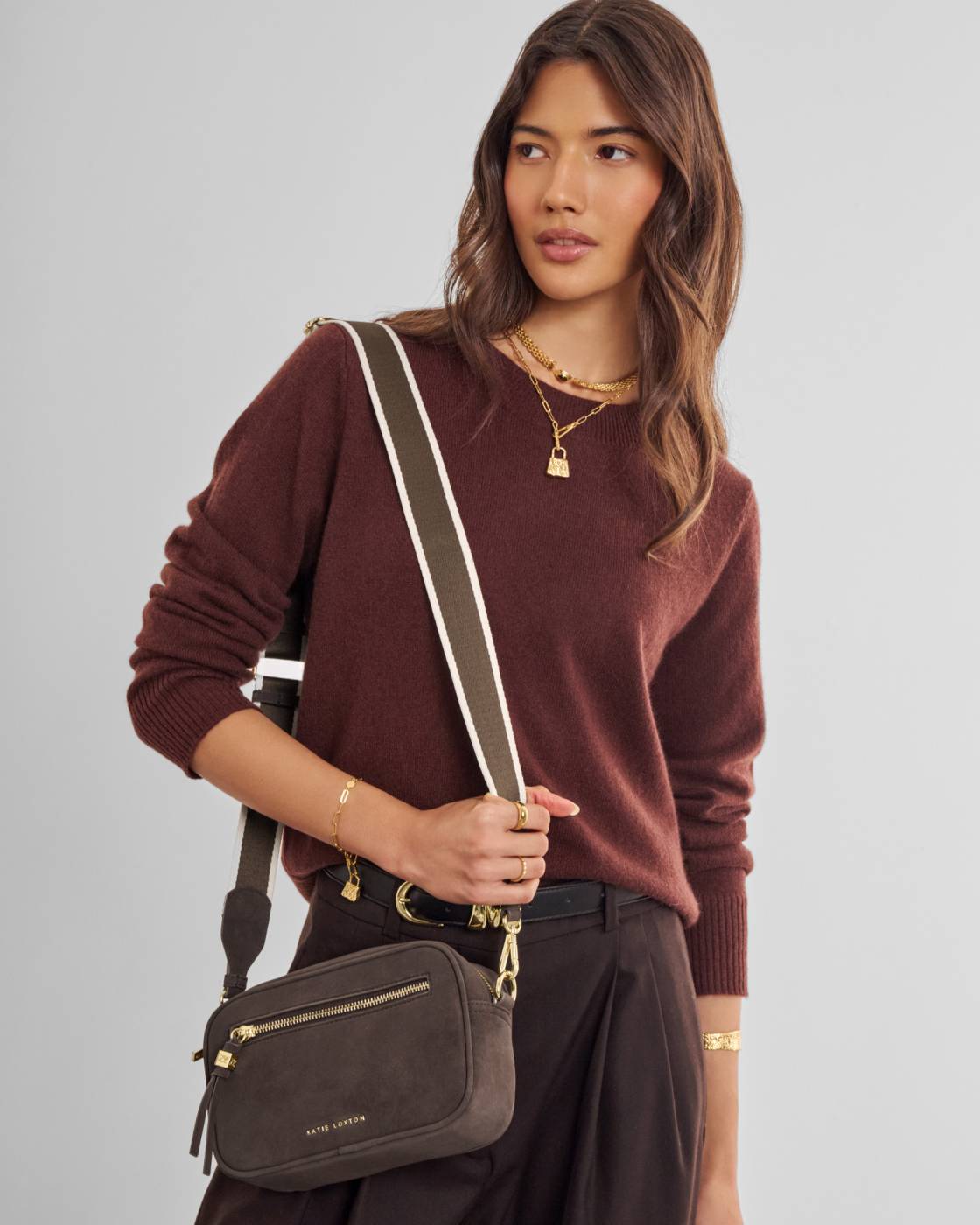 A woman wearing a brown knit sweater and dark trousers carries a dark brown crossbody bag with a wide olive green and white striped strap. The bag has gold-tone hardware and a small gold logo on the front, photographed against a solid light gray background.