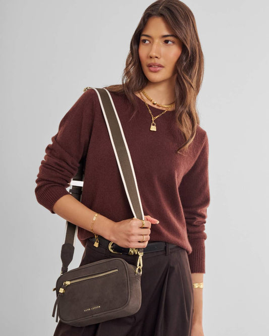 A woman wearing a brown knit sweater and dark trousers carries a dark brown crossbody bag with a wide olive green and white striped strap. The bag has gold-tone hardware and a small gold logo on the front, photographed against a solid light gray background.