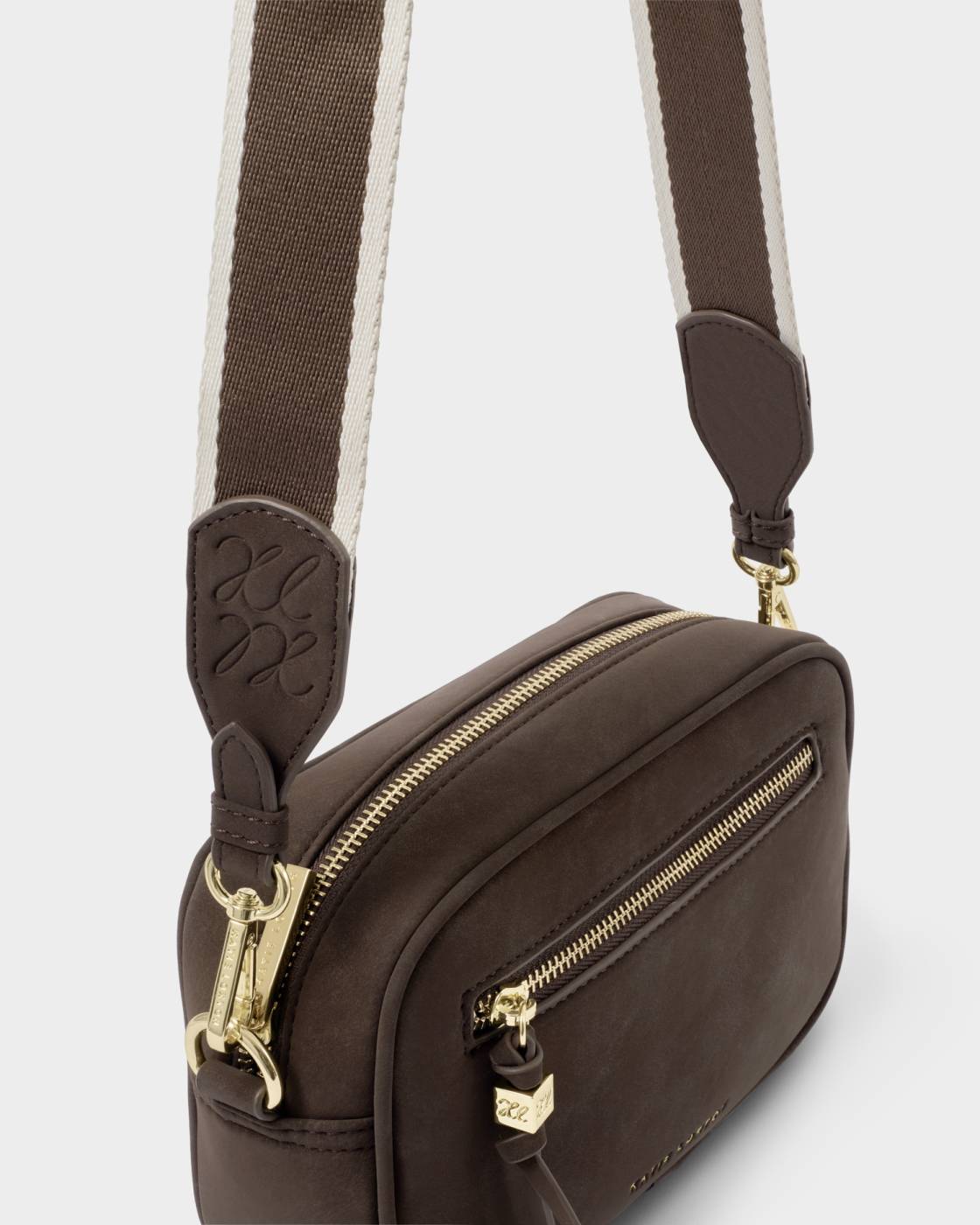 A dark brown crossbody bag with a matching wide woven strap featuring a cream stripe, gold-tone hardware, and a small tassel zipper pull on a light gray background.