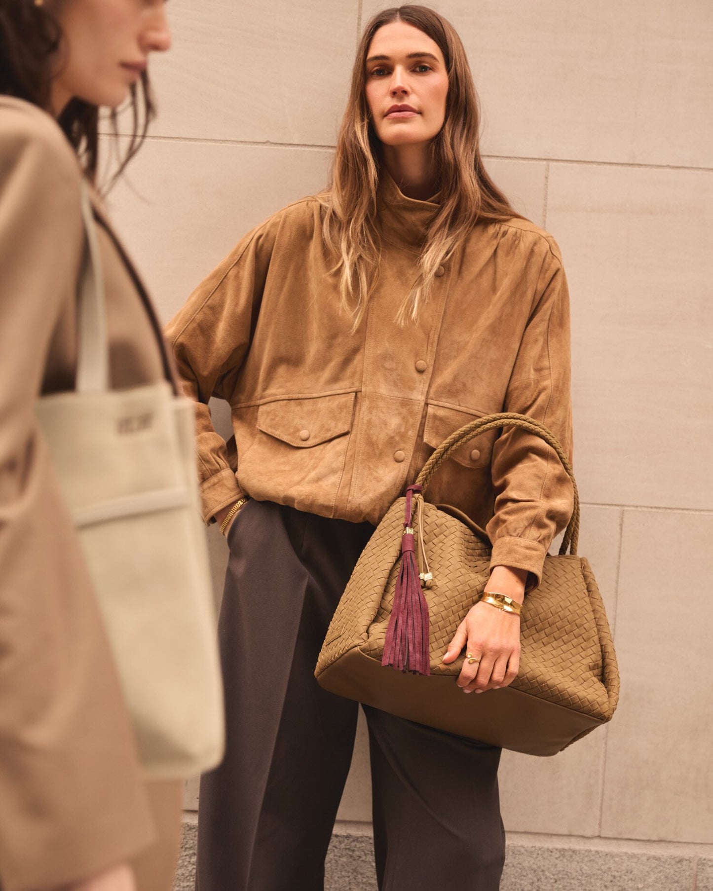 Woman holding a brown handbag with a tassel, wearing a beige jacket and dark pants, against a neutral wall.