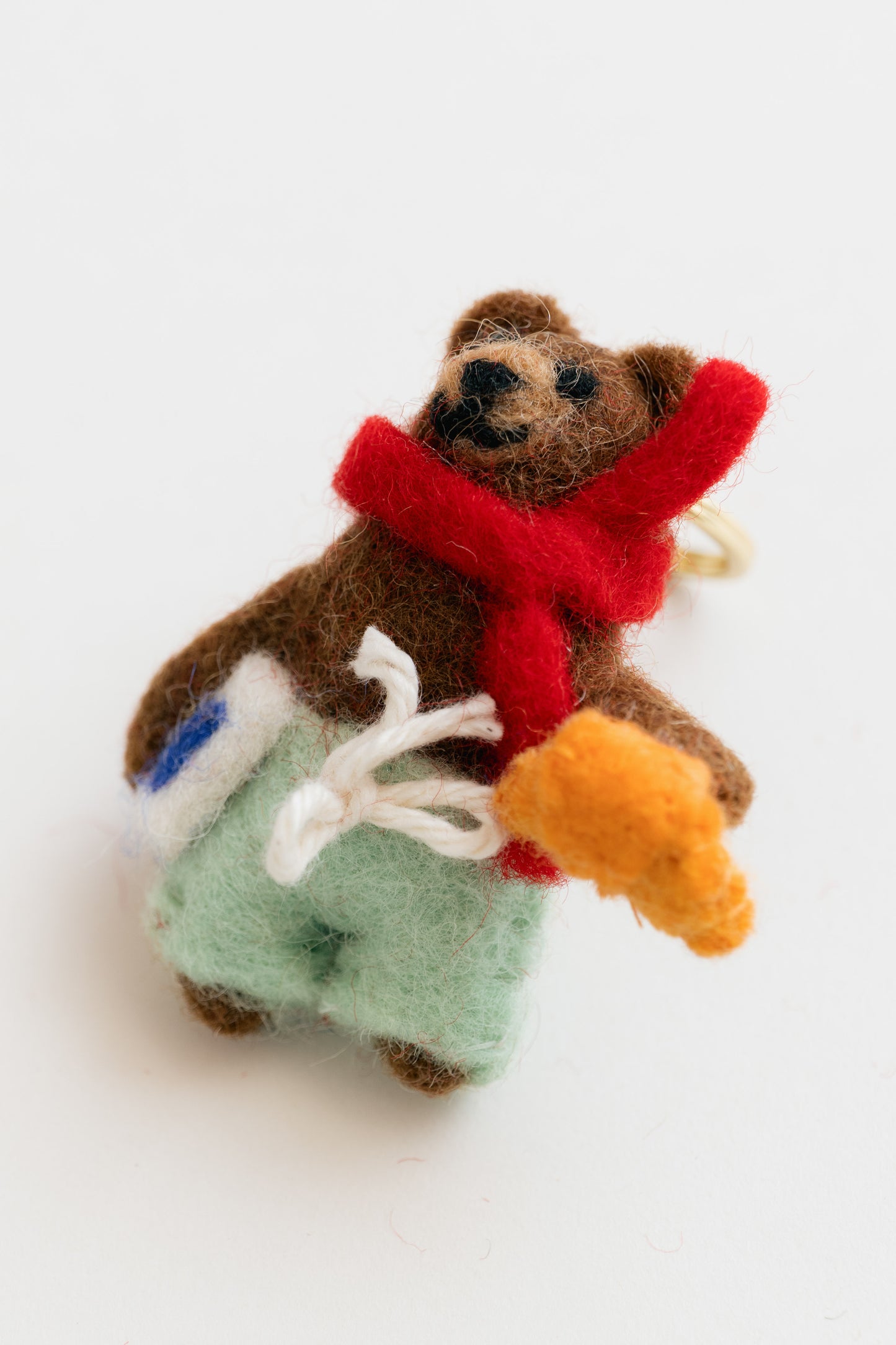 Small felted bear keychain with a red scarf, green pants, and an orange carrot on a white background