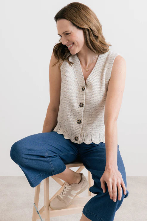 Woman wearing a beige sleeveless sweater and blue pants sitting on a stool.