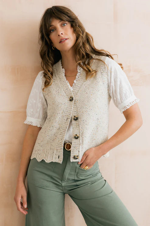 Woman wearing a white lace cardigan over a white shirt with green pants against a beige wall.