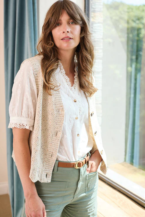 Woman wearing a white blouse, beige cardigan, and green pants in a bright room.