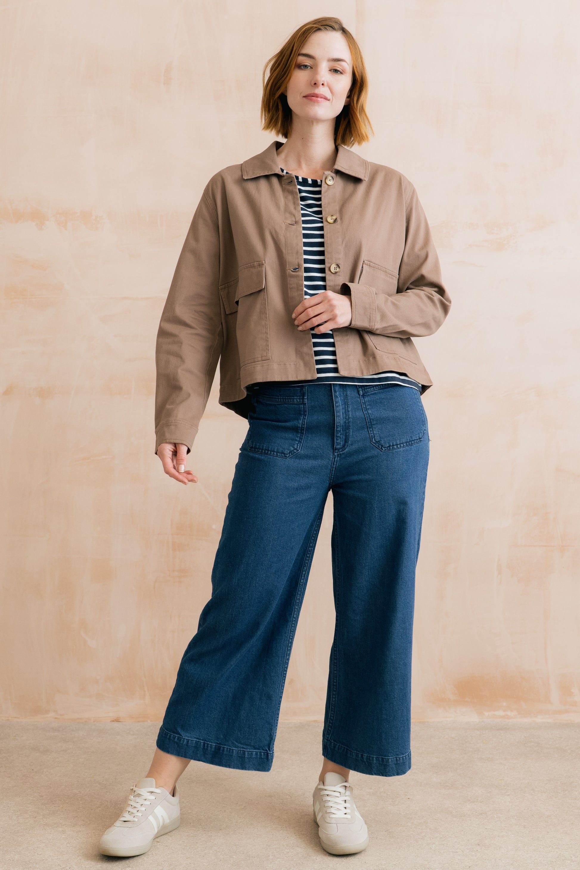 Woman wearing a beige jacket, striped shirt, and blue jeans against a beige background