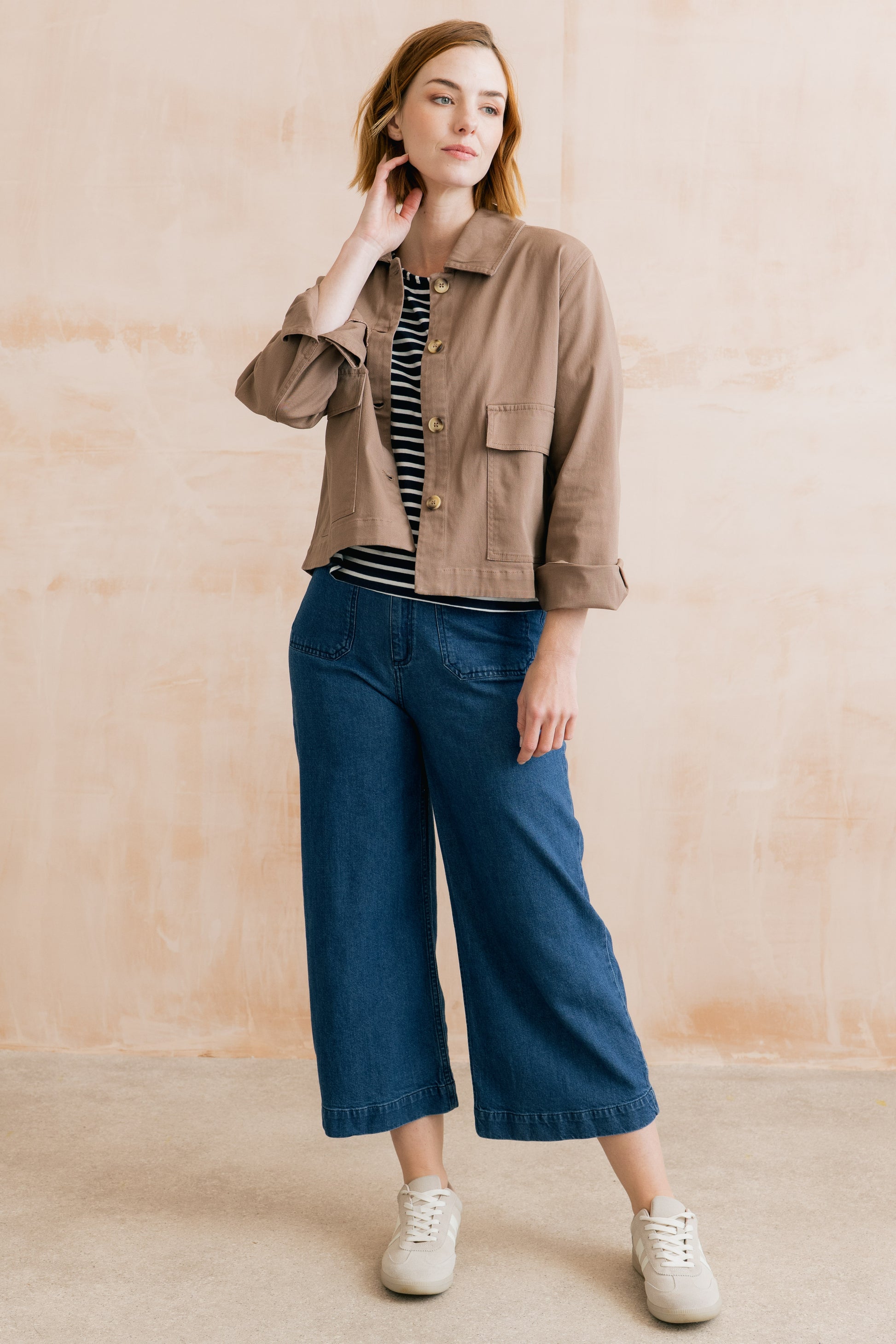 Woman wearing a brown jacket, striped shirt, blue jeans, and white sneakers against a beige wall.