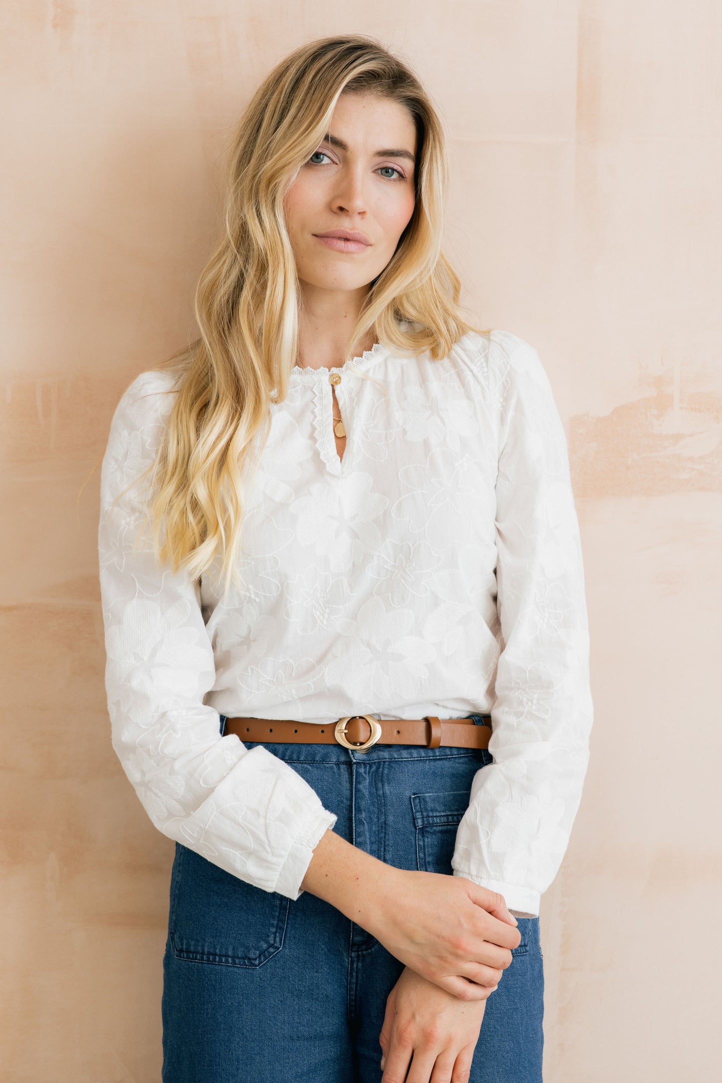 Woman wearing a white blouse with long sleeves and blue jeans against a beige background