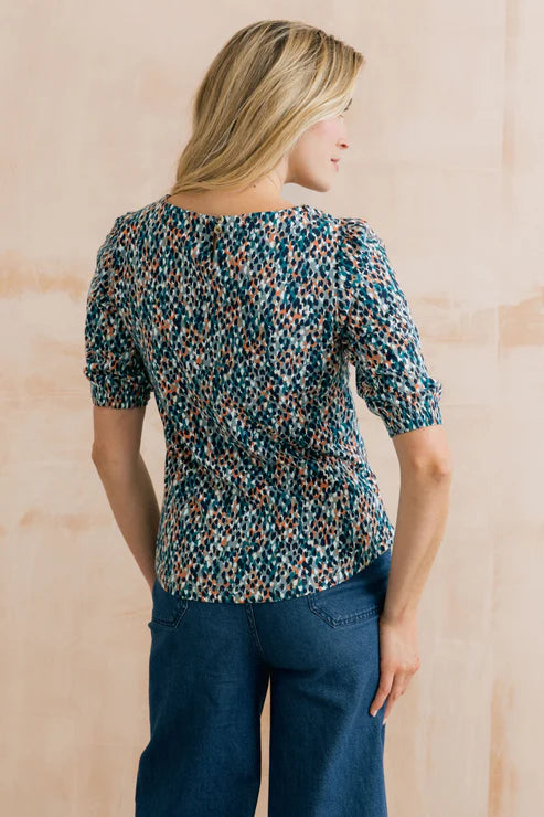 Woman wearing a patterned blouse and blue jeans against a wooden background