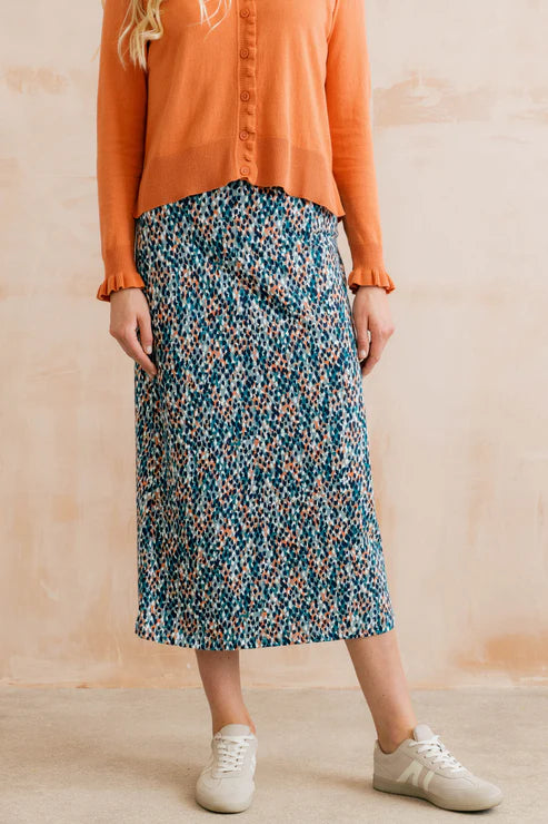 Person wearing an orange shirt and a patterned blue skirt against a beige background