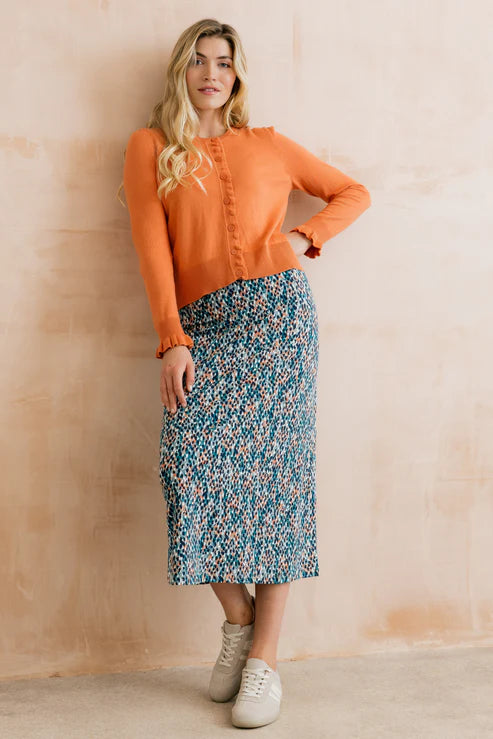 Woman wearing an orange sweater and patterned skirt against a beige wall