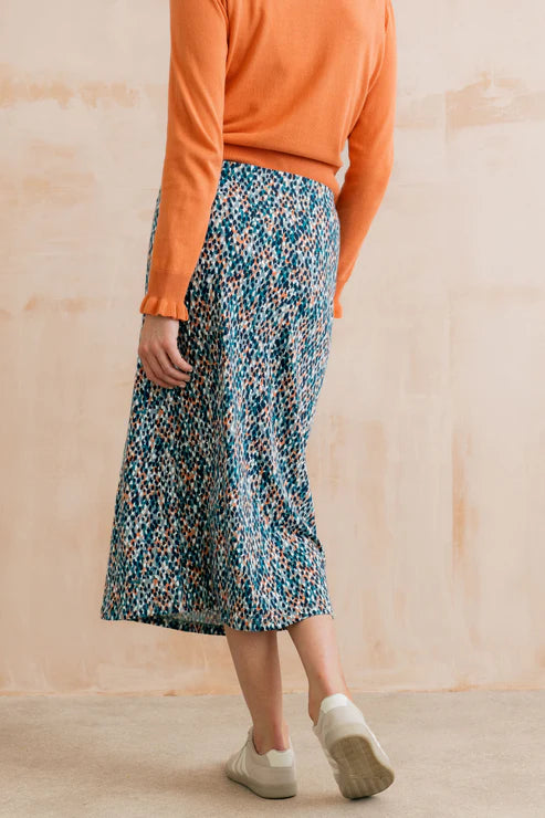 Person wearing an orange top and patterned pants against a beige wall.