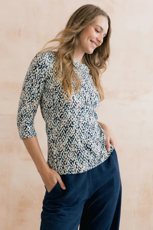 Woman wearing a patterned top and navy pants against a light wooden background