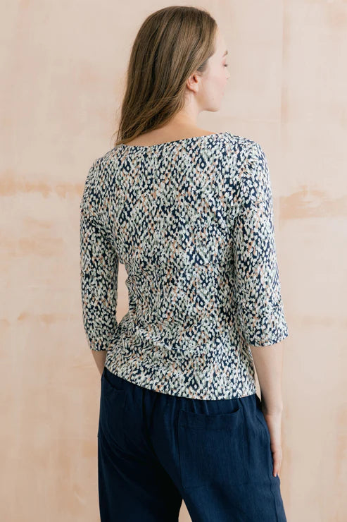 Woman wearing a patterned blouse and dark pants against a beige wall