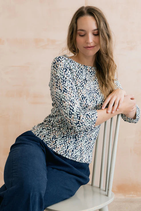 Woman sitting on a chair wearing a patterned blouse and blue pants against a beige wall.