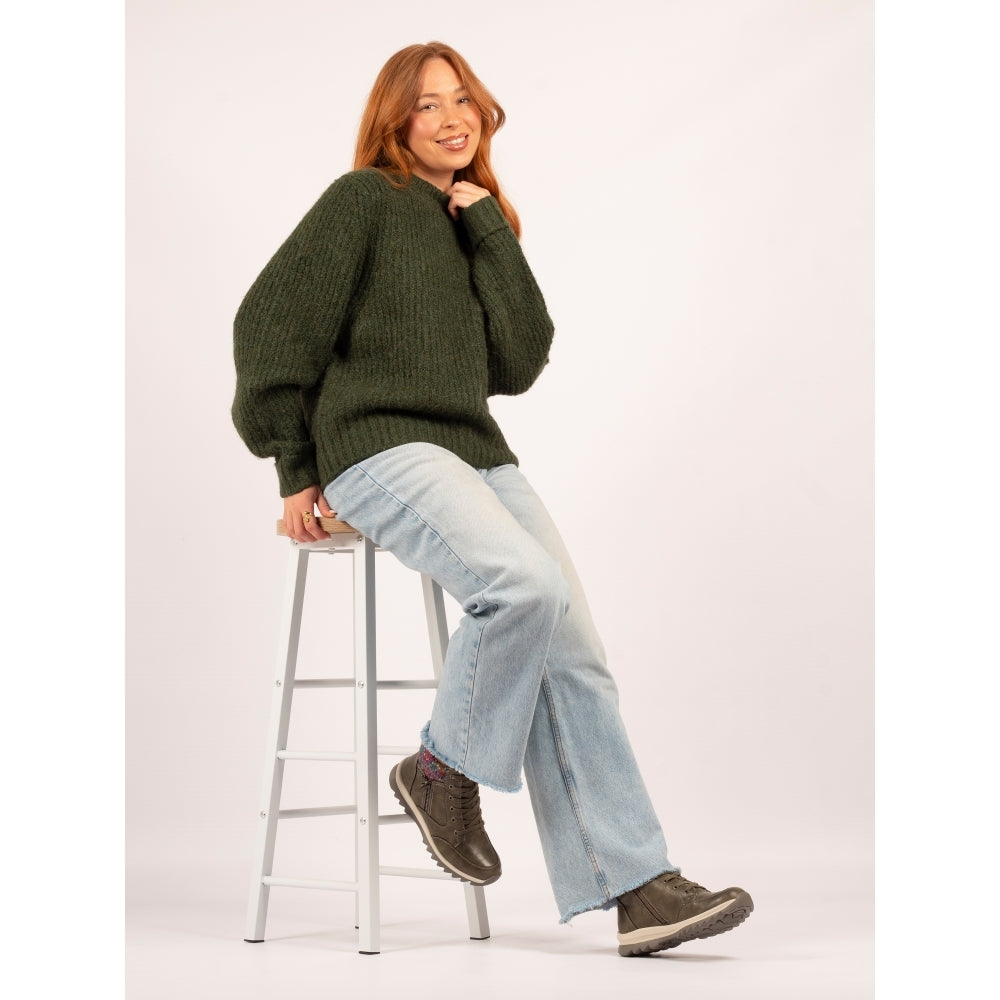 Person wearing a green sweater and light blue jeans sitting on a stool against a white background