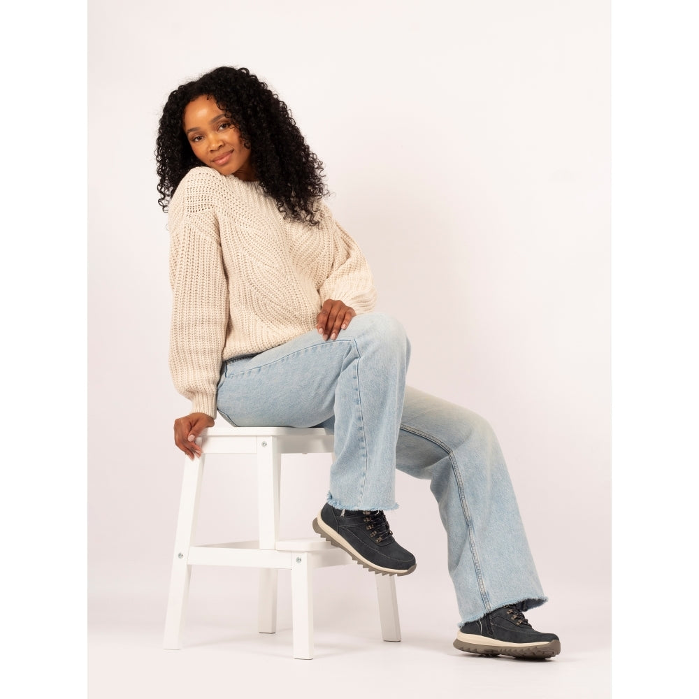 Woman sitting on a stool wearing a cream sweater, light blue jeans, and black shoes against a white background