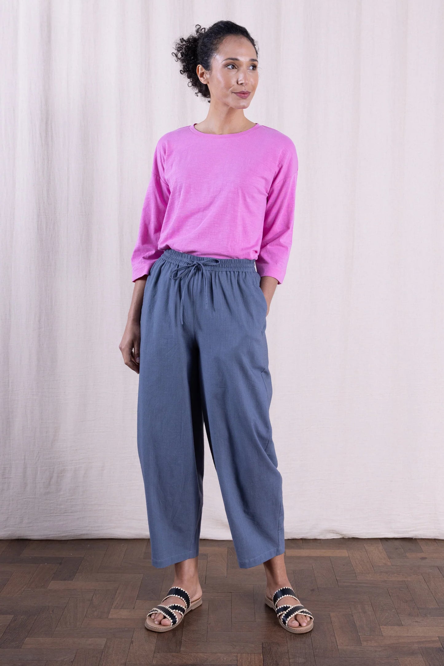 Woman wearing a pink top and gray pants standing against a white curtain.