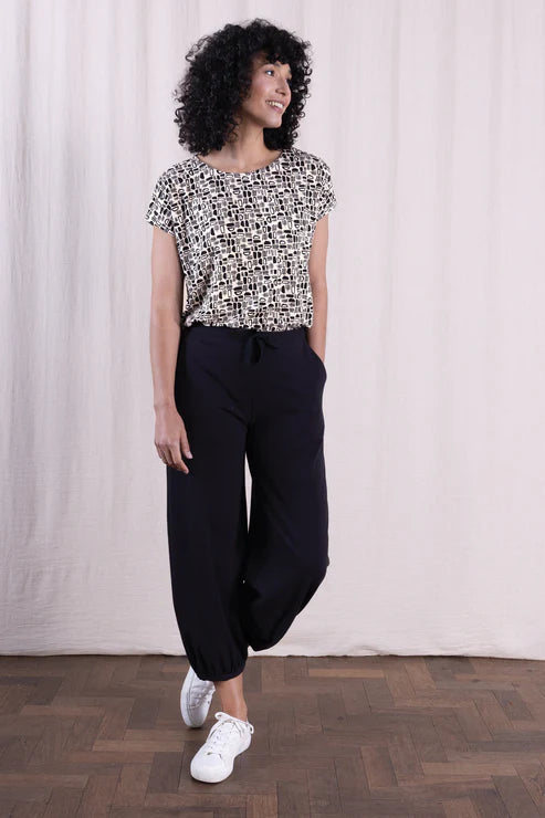Woman wearing a patterned top and black pants standing against a white curtain.