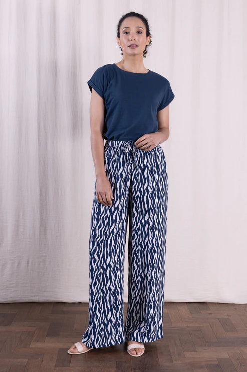 Woman wearing a navy blue top and patterned pants standing against a white curtain.