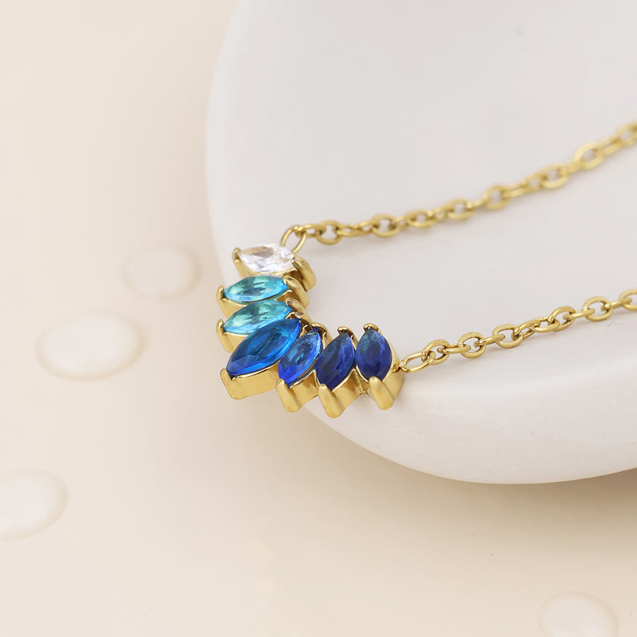 Gold chain necklace with a crescent-shaped pendant featuring marquise-cut blue and royal blue stones, a clear round stone, and a small white stone, displayed on a white curved surface with water droplets.