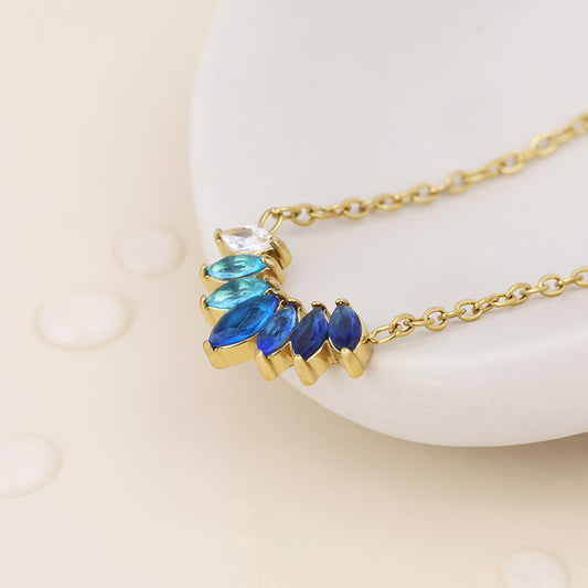 Gold chain necklace with a crescent-shaped pendant featuring marquise-cut blue and royal blue stones, a clear round stone, and a small white stone, displayed on a white curved surface with water droplets.