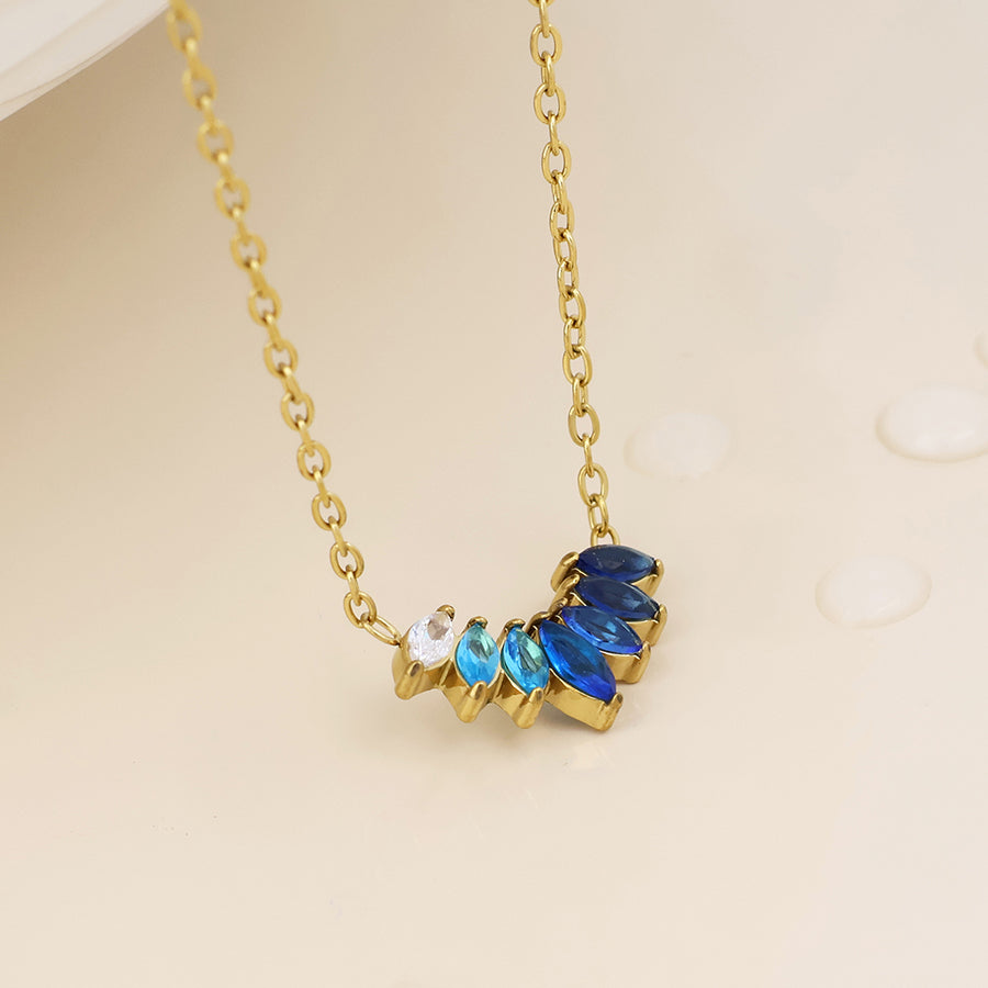 A gold chain necklace with a curved pendant featuring blue and light blue teardrop-shaped stones and a small clear round stone, displayed on a light beige background.
