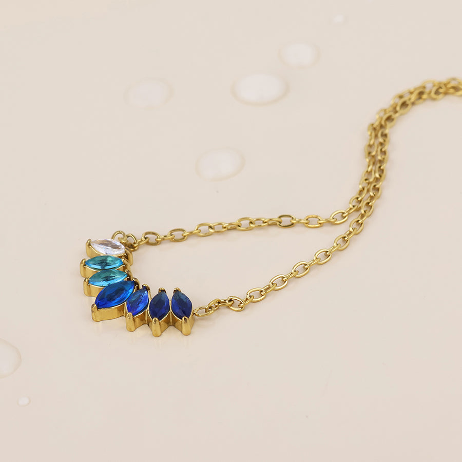 A gold chain necklace with a curved cluster of marquise-cut gemstones in white, light blue, royal blue, and dark blue, displayed on a light beige background with subtle water droplets.