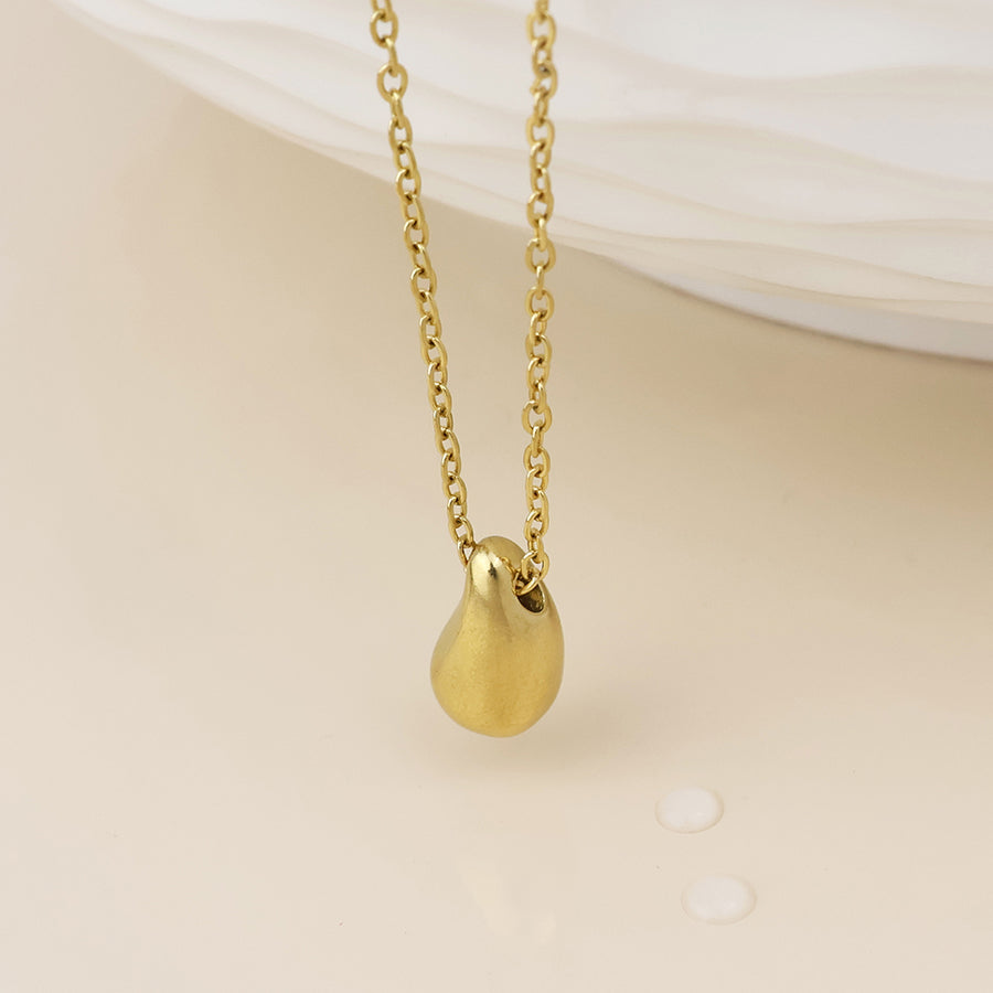 A gold chain necklace with a smooth, teardrop-shaped gold pendant, displayed against a soft beige background with a white textured surface in the upper right.