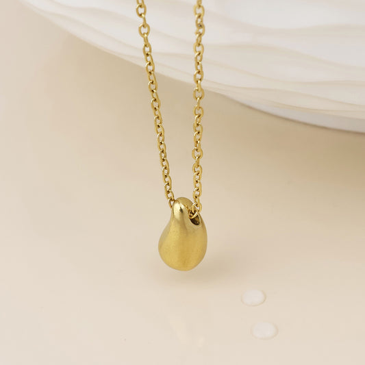 A gold chain necklace with a smooth, teardrop-shaped gold pendant, displayed against a soft beige background with a white textured surface in the upper right.