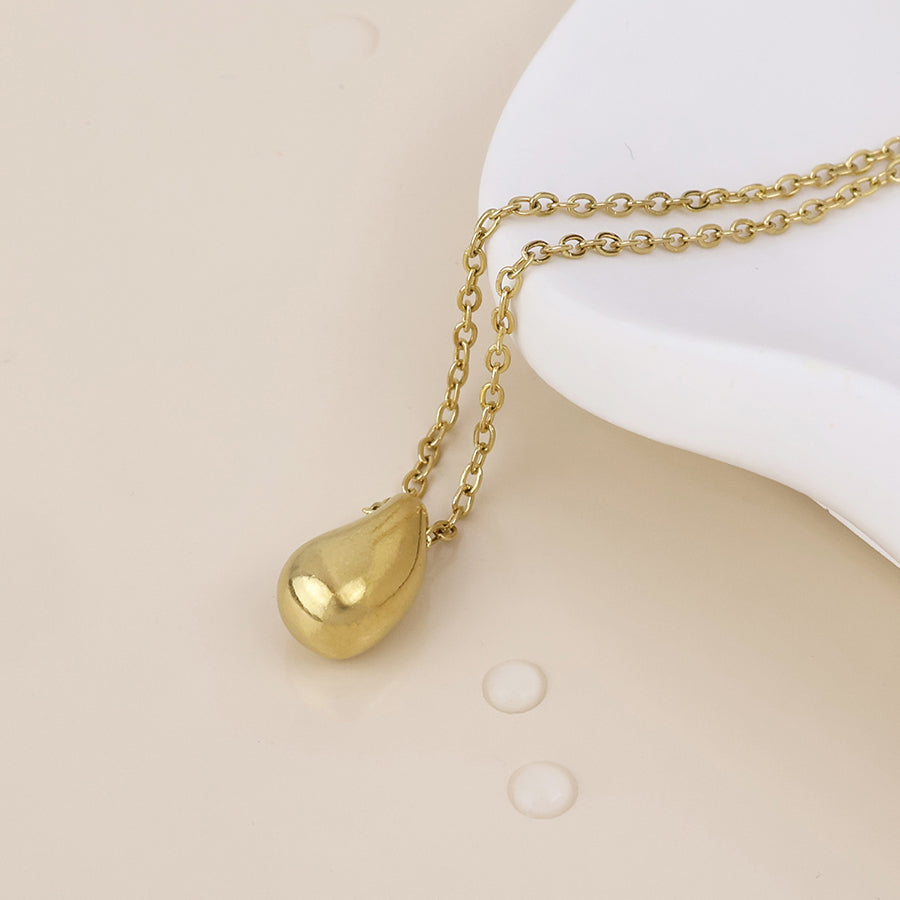 A gold teardrop pendant necklace on a delicate gold chain, styled on a white ceramic dish against a warm beige background with water droplets.