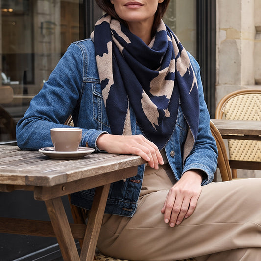 A woman seated at an outdoor cafe table wearing a navy and beige patterned scarf, a blue denim jacket, and beige trousers, with a cup of coffee on the table.