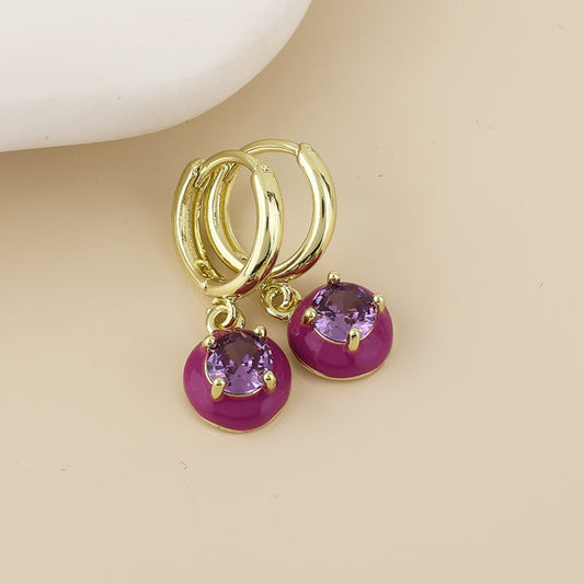 A pair of gold huggie hoop earrings with round purple gemstone charms, displayed on a light beige surface.