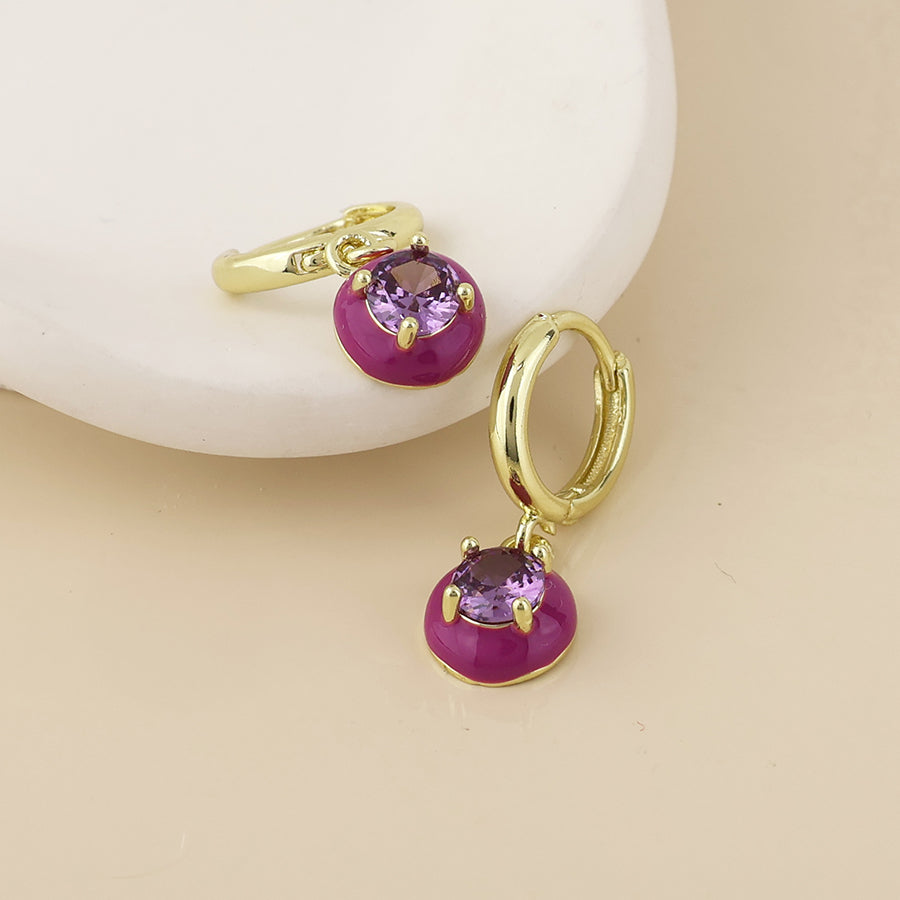 A pair of gold huggie hoop earrings with dangling purple oval charms, displayed on a light beige surface with a white curved prop.