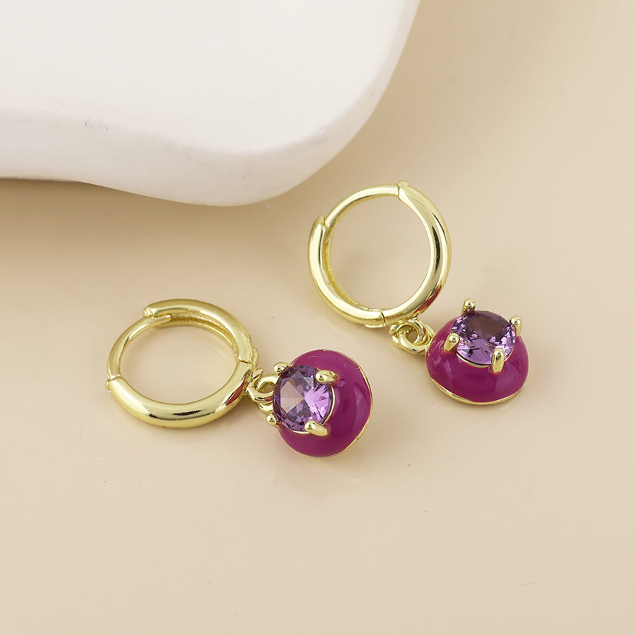 A pair of gold hoop earrings with round magenta-purple gemstone charms, displayed on a light beige surface with a white ceramic prop in the background.