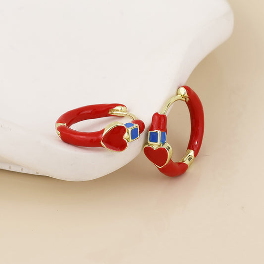 A pair of gold hoop earrings with red enamel sections, a small red heart, and a blue square accent, displayed on a white curved stand against a light beige background.