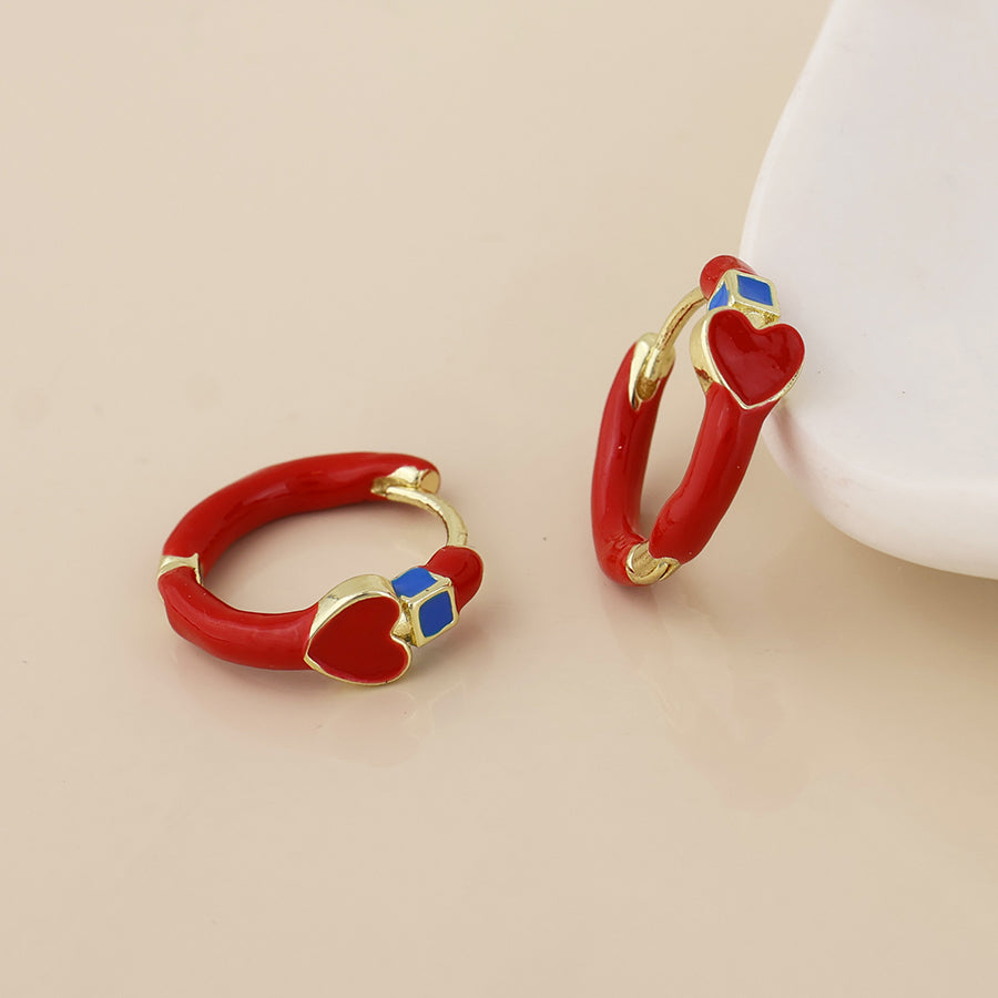 A pair of small hoop earrings with glossy red enamel, gold-tone metal borders, and a small blue square accent near the top, displayed on a light beige surface with a white display stand partially visible.