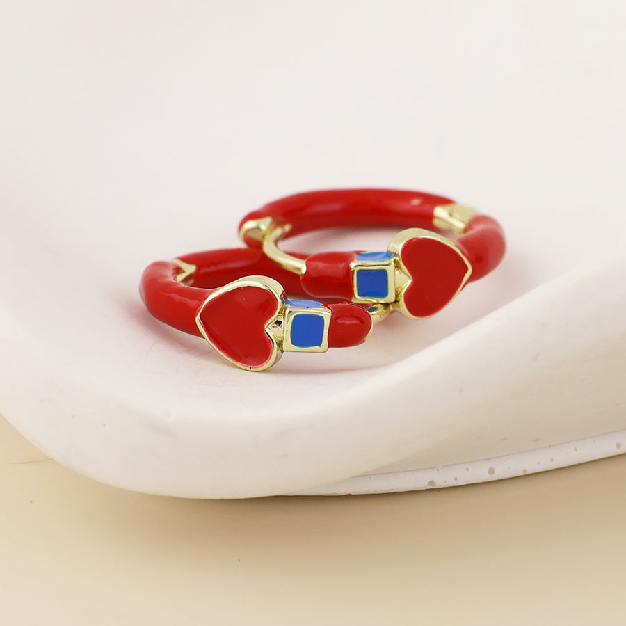 A pair of red enamel rings with gold-plated edges, featuring heart-shaped details and small blue square accents, displayed on a white ceramic dish.