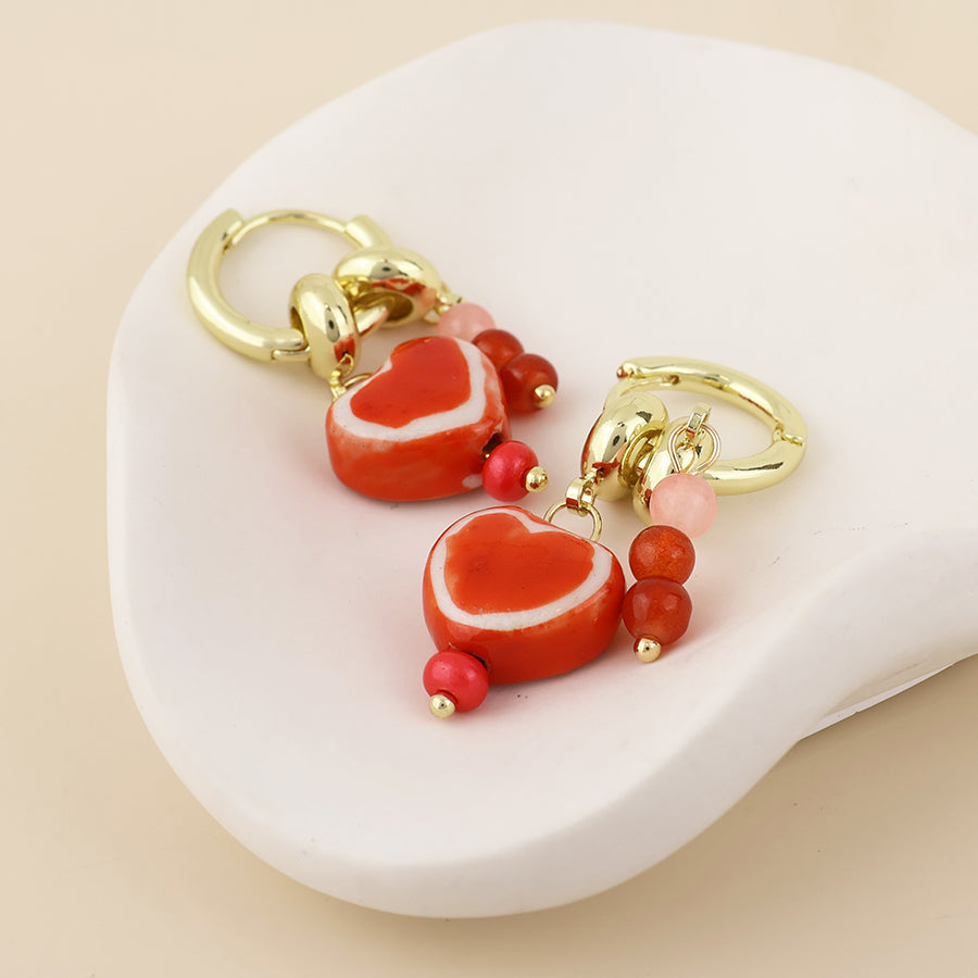 A pair of gold-tone dangle earrings with red heart-shaped beads outlined in white, accented with small pink and red beads, displayed on a white ceramic dish against a light beige background.