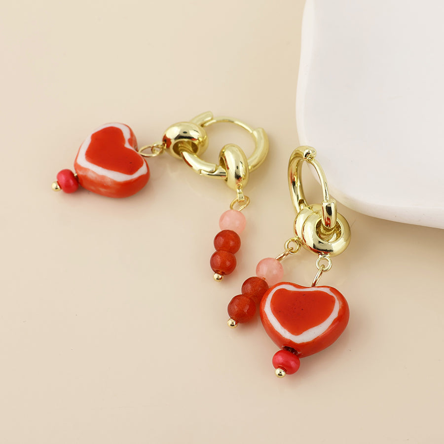 A pair of gold-tone dangle earrings with red and white heart-shaped beads, pink beads, and small red beads, displayed on a light beige background.