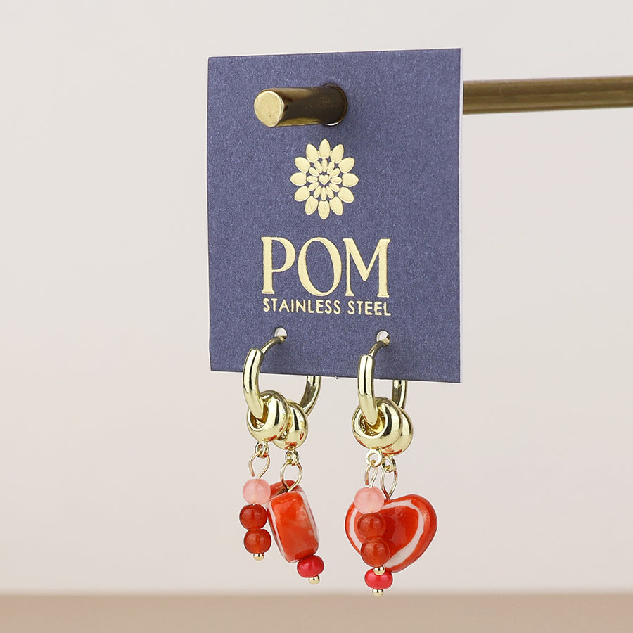 A pair of POM stainless steel dangle earrings with red and orange heart-shaped beads, hanging from gold-tone hoops on a dark blue display card.